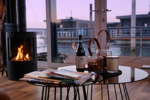 A cozy room with a wood-burning stove, a bottle of wine, a candle, and a glass of water on a table, with a view of buildings and water outside.