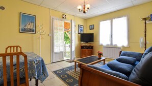 Un salon comprenant un canapé, une table, un téléviseur et un fauteuil. Une porte donne accès à un balcon d’où l’on peut admirer la verdure.
