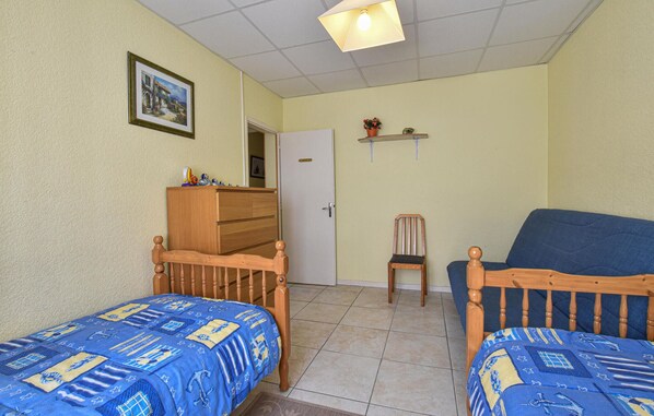 A room with two wooden beds with blue bedding, a wooden dresser, a chair, a blue sofa, a door, and a shelf with flowers.