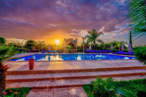 Relax by the serene pool surrounded by lush gardens and palm trees, with lounge chairs and umbrellas for a peaceful retreat.
