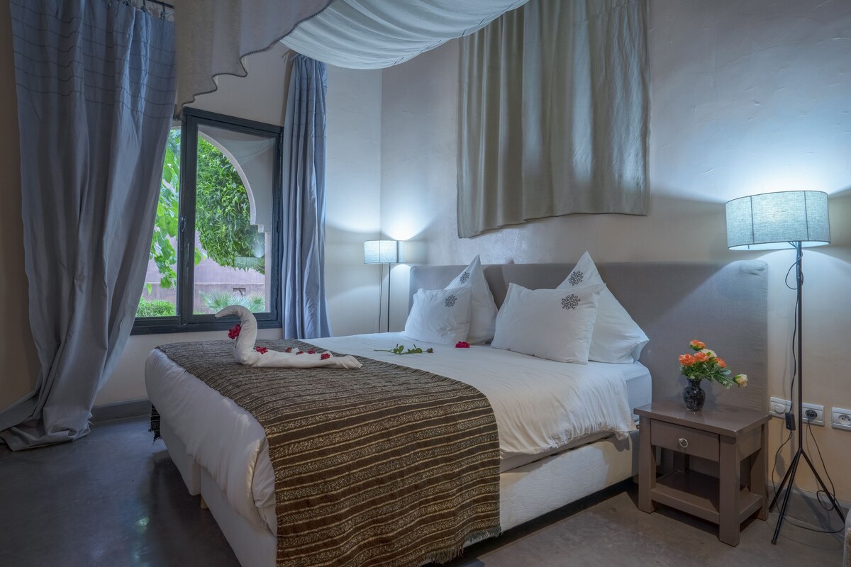 A hotel room with a large bed, a bedside table with a lamp, and a window with curtains.