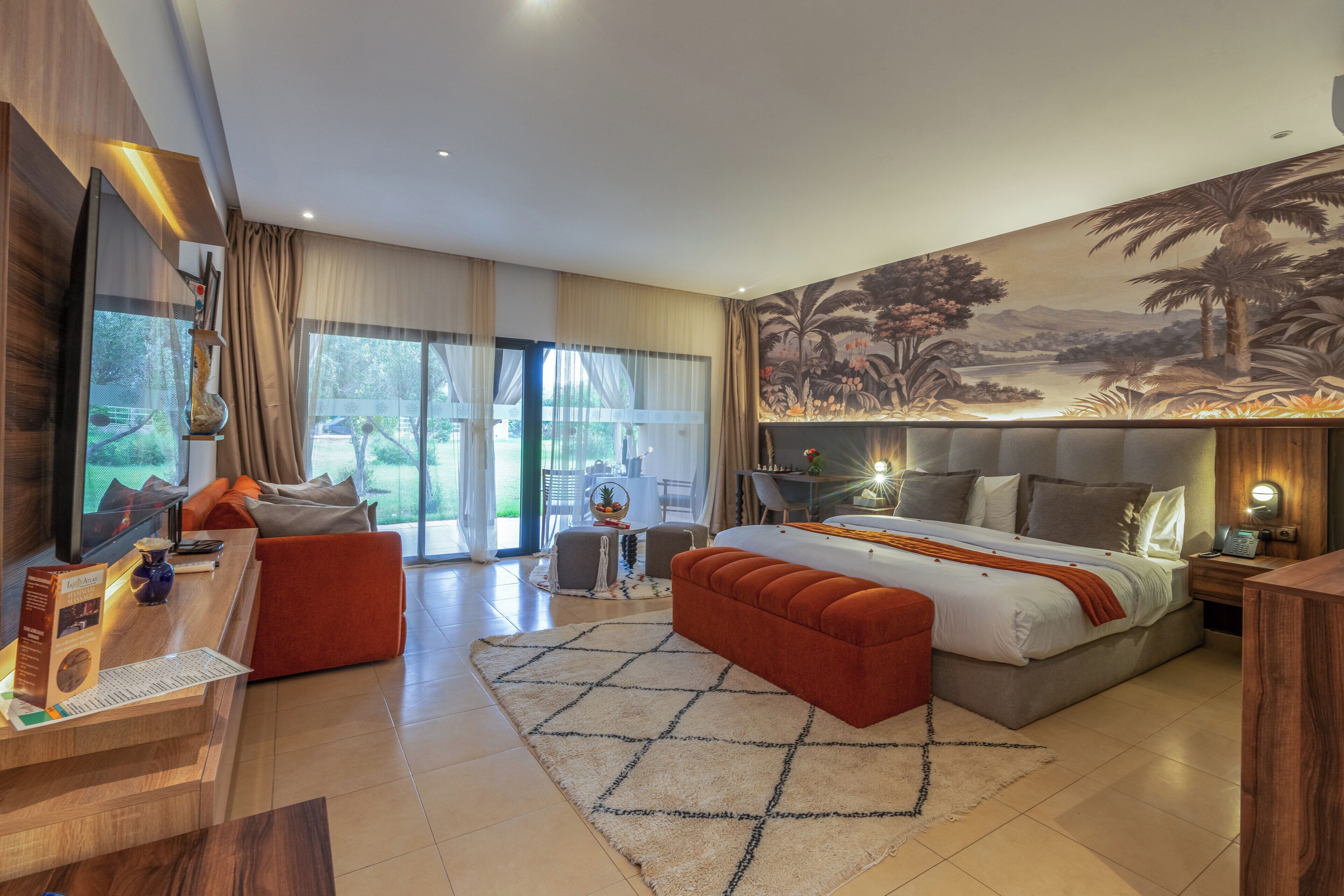 Una habitación de hotel moderna con una cama grande, un escritorio con computadora, una zona de estar con sofá y sillones, y un mural de paisaje tropical en la pared.