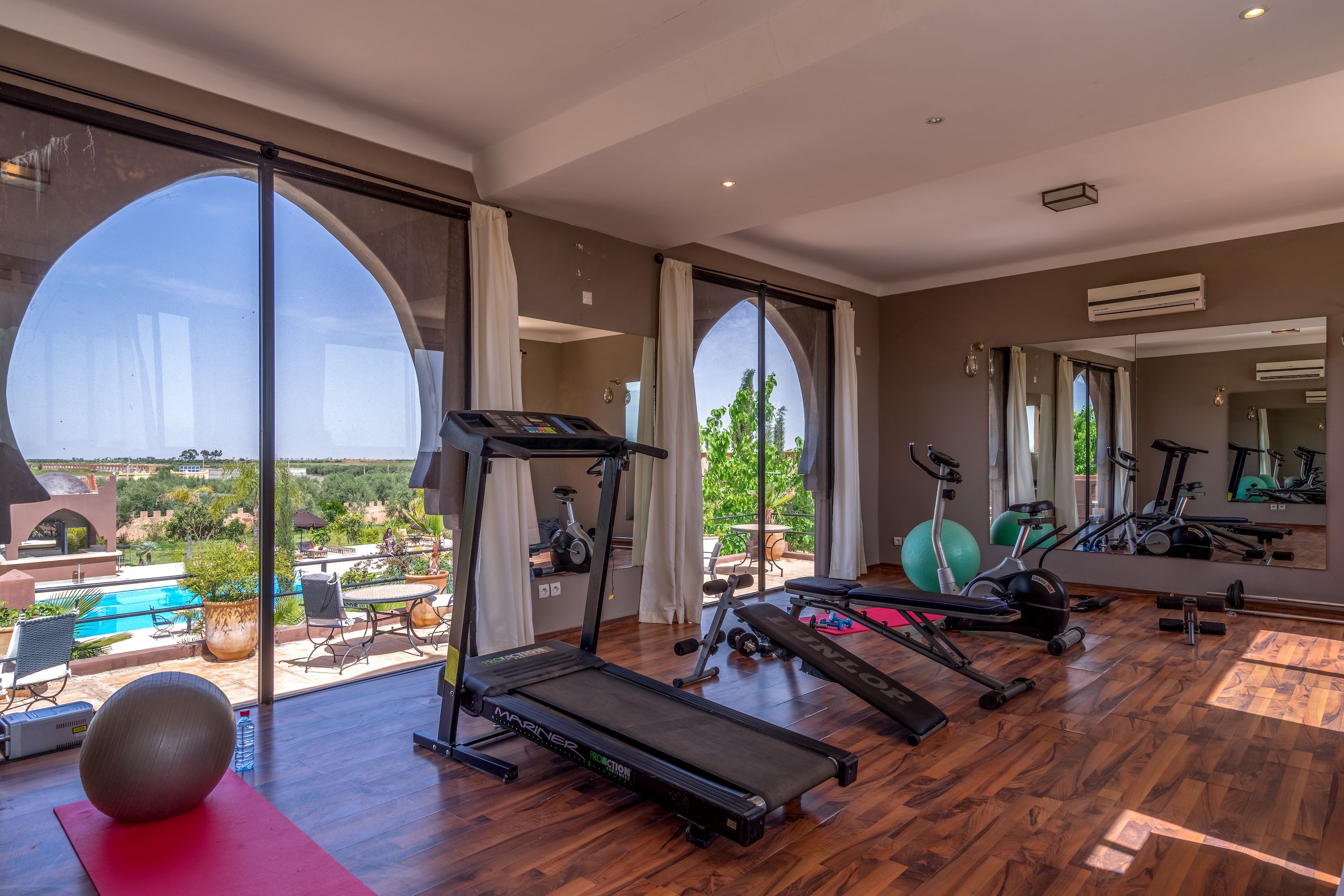 Un gimnasio amplio en casa con cinta de correr, pelotas de ejercicio y pesas. Grandes ventanales ofrecen vistas a una piscina y follaje.