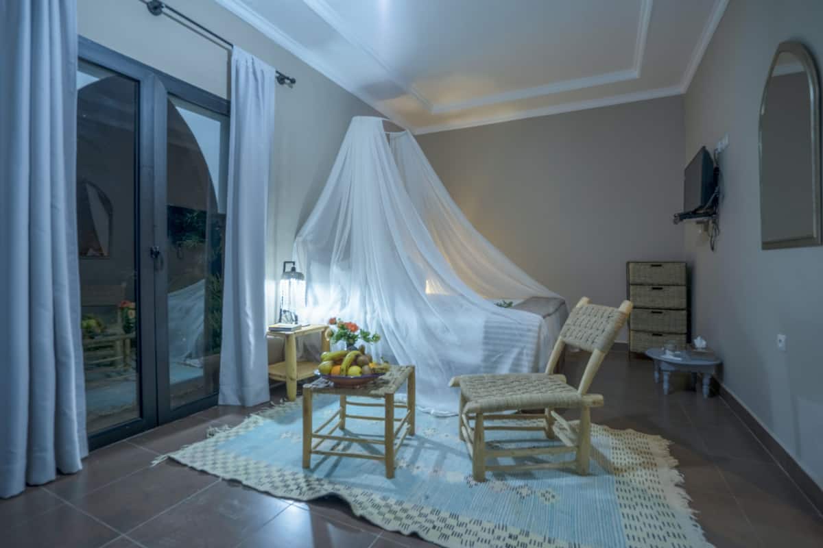 Een slaapkamer met een hemelbed, een klein tafeltje met fruit, een rieten stoel en uitzicht door een glazen deur.