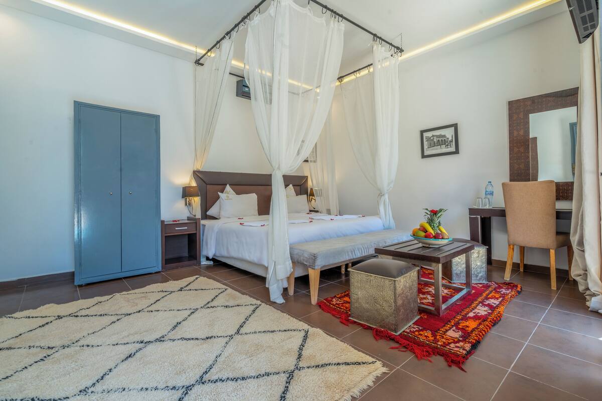 Een slaapkamer met een hemelbed, een bureau met stoel, een kledingkast en een kleine tafel met een fruitschaal.