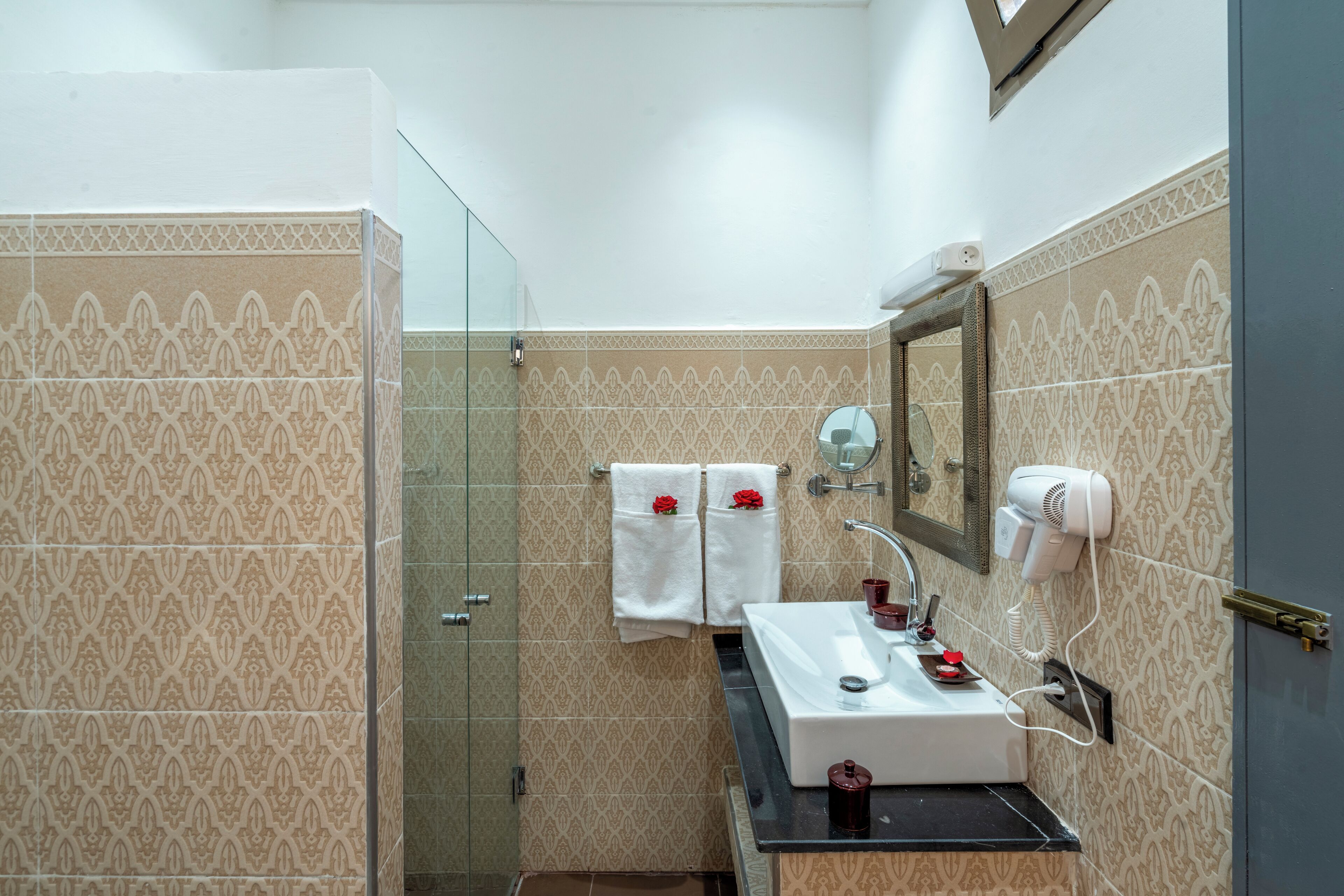 A bathroom with patterned tiles, a glass shower enclosure, a sink with a mirror, and a hairdryer on the wall.