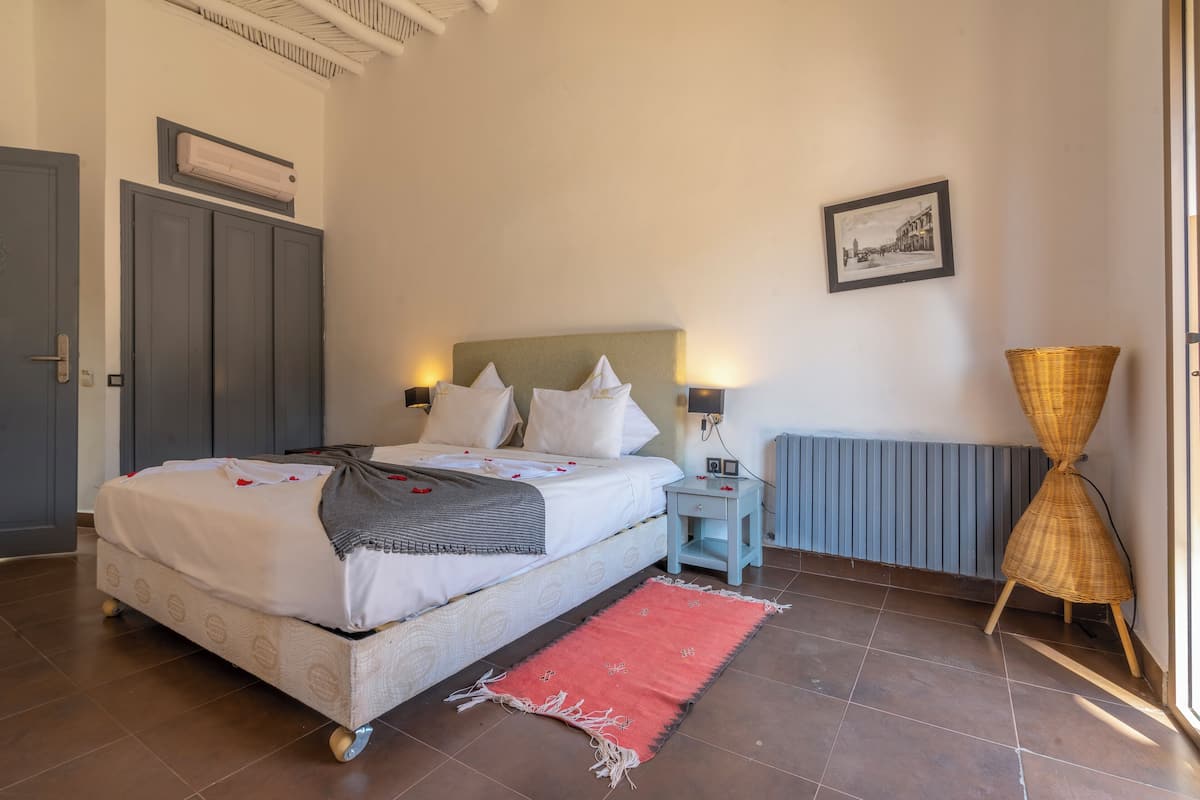 Een slaapkamer met een bed, nachtkastje, lamp en een rieten stoel.
