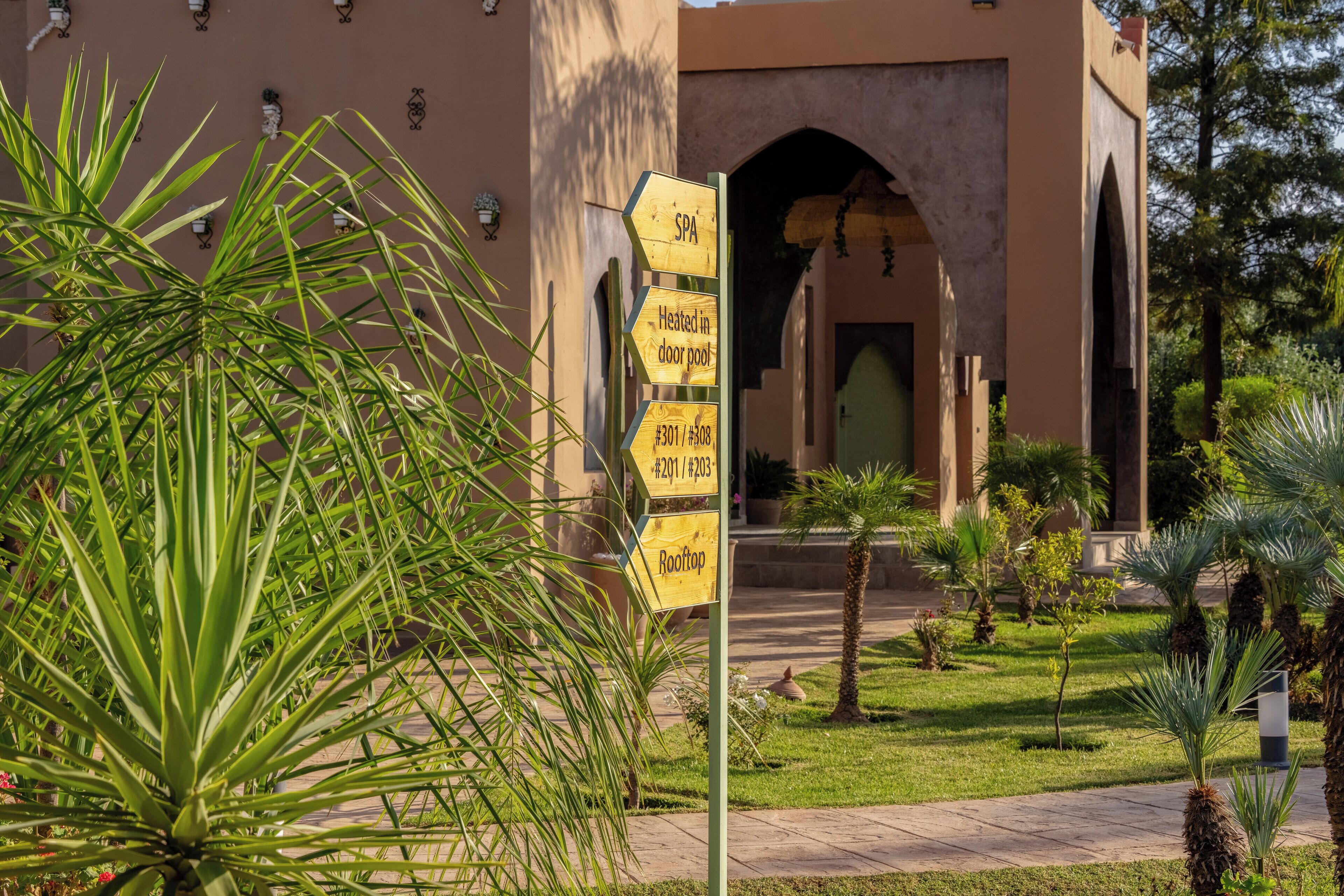 A signpost with directions to a spa, heated door pool, and rooftop area, located in a courtyard with palm trees and a building with arches in the background.