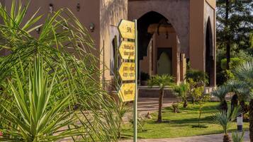A signpost with directions to a spa, heated door pool, and rooftop area, located in a courtyard with palm trees and a building with arches in the background.