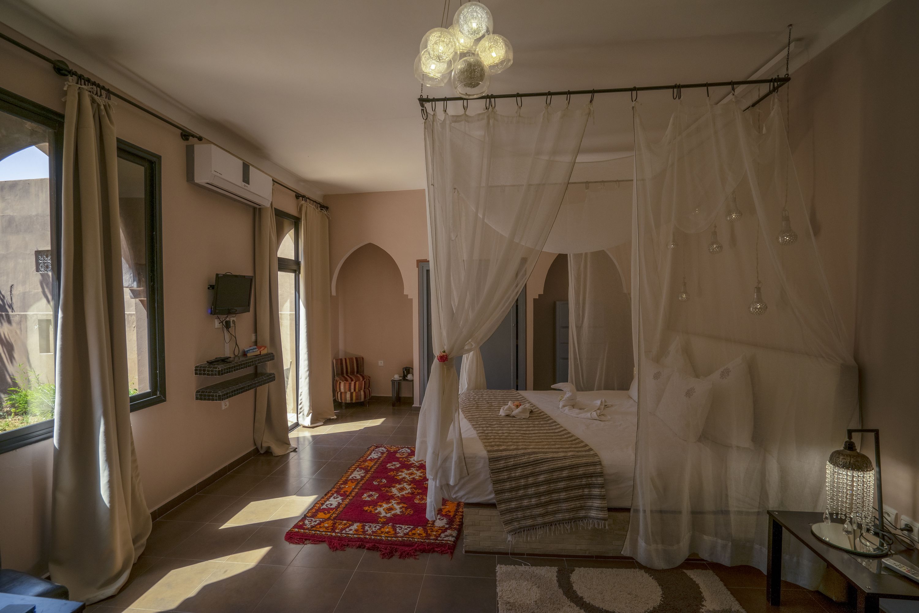 Un dormitorio con cama con dosel, un tapete rojo estampado y una mesita de noche con lámpara.