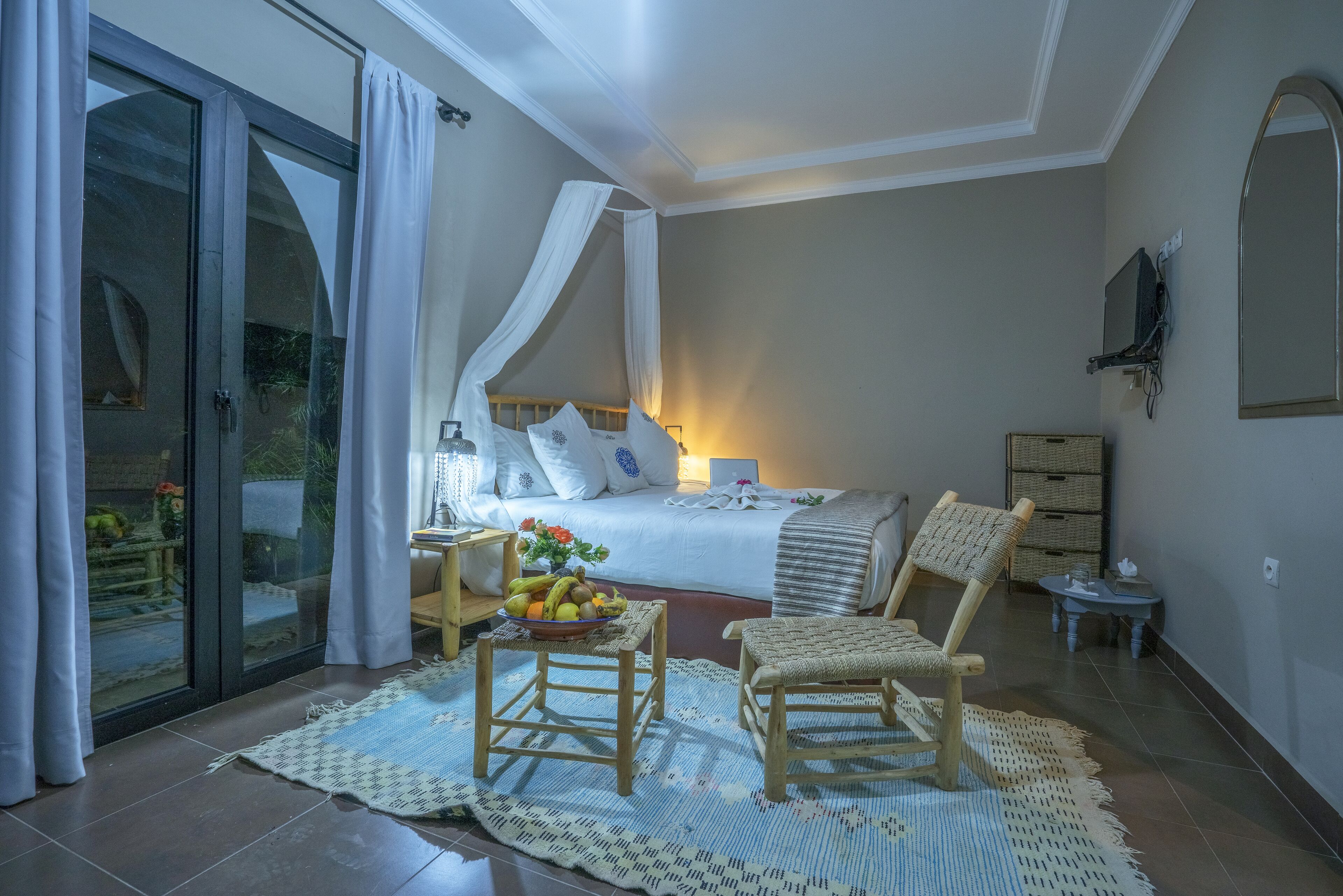 Un dormitorio con cama, una mesita de noche con lámpara, una silla, una pequeña mesa con frutas y un amplio ventanal con cortinas.