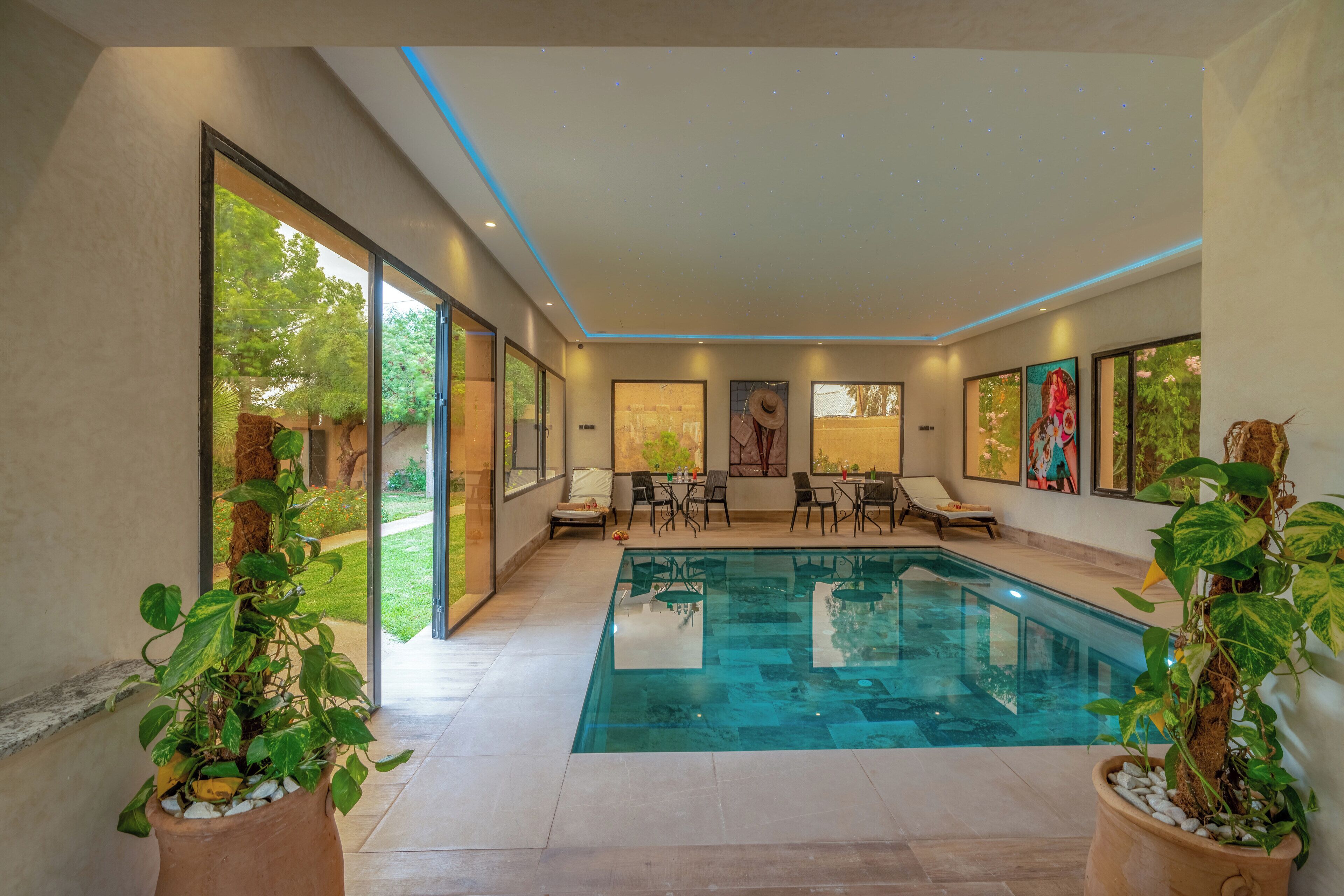 Un área de piscina interior moderna con una piscina de vidrio transparente, rodeada de sillones y mesas, y amplias ventanas con vistas al exterior.