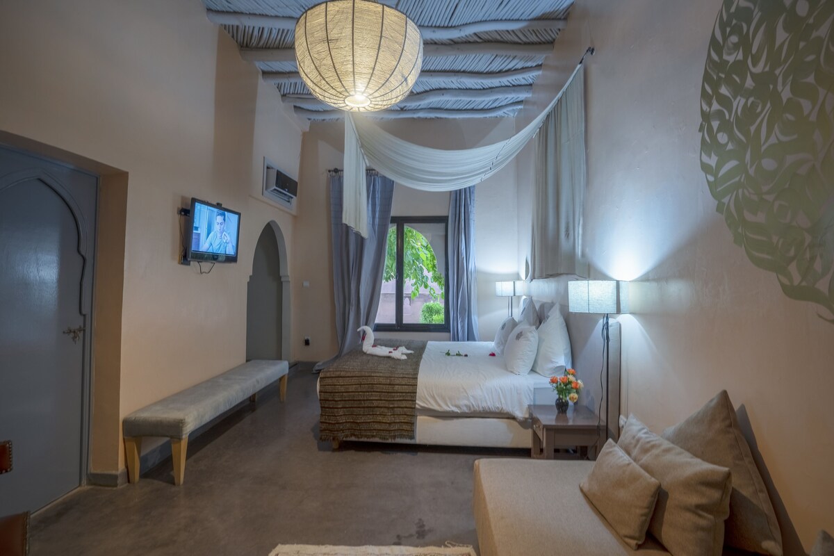 Een slaapkamer met een bed, een bankje, nachtkastje, lamp en een televisie aan de muur.