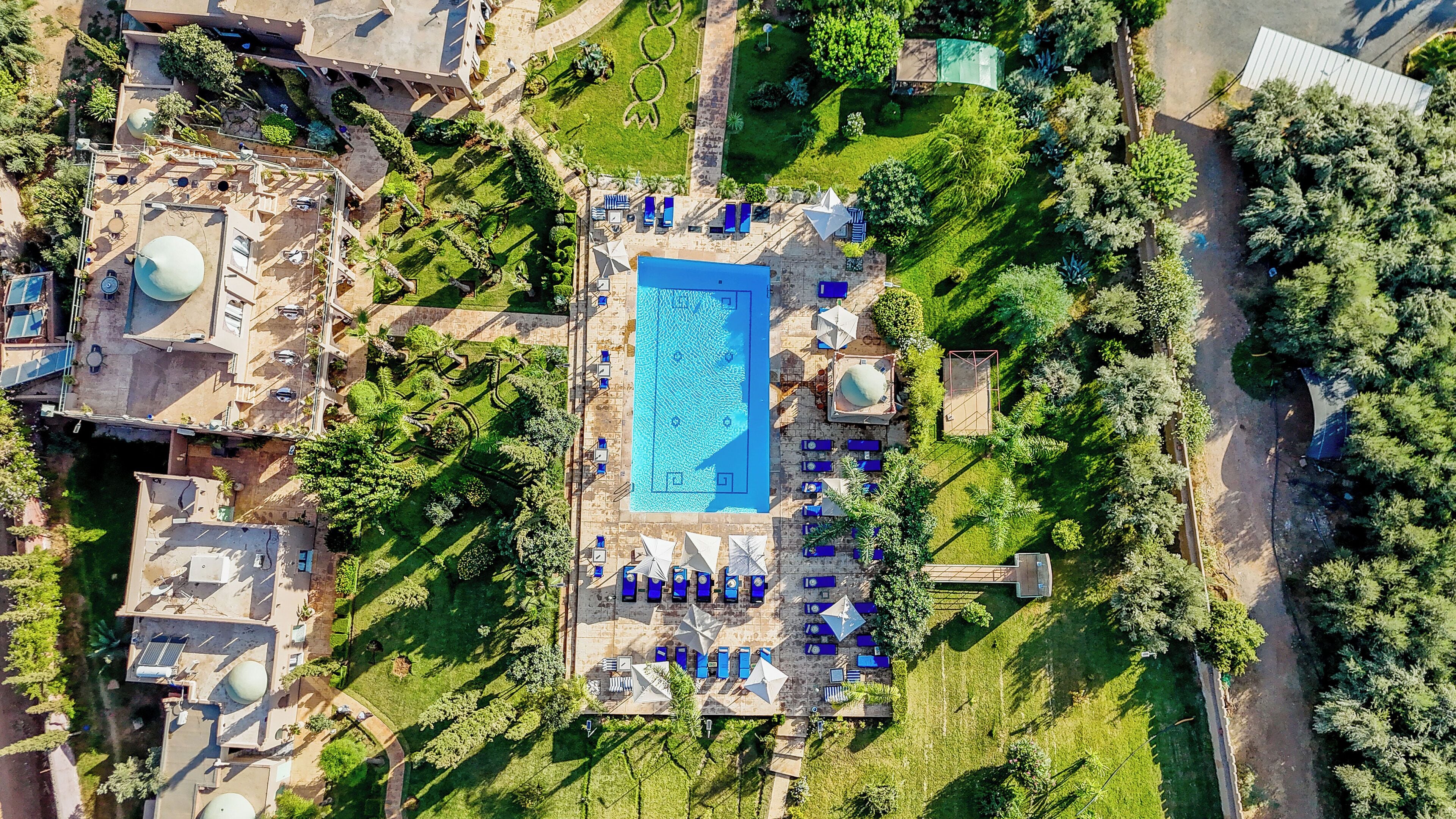 Luchtbeeld van een resort met een centraal zwembad, omringd door ligbedden en parasols, een aangelegde tuin en een gebouw met een koepelstructuur.