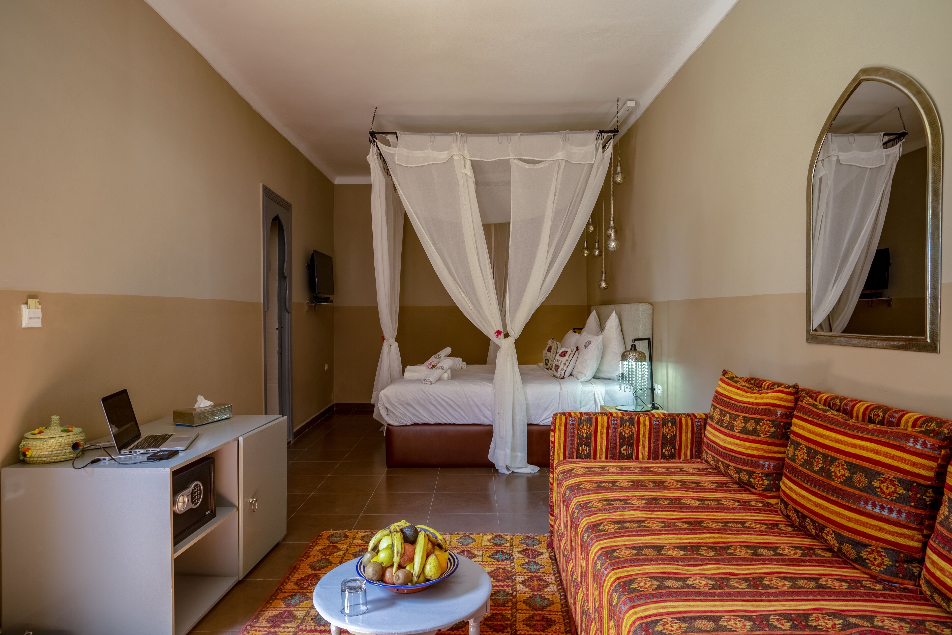 Un dormitorio con una cama, un sofá, un escritorio con una computadora portátil, una mesita de noche con un cuenco de fruta y una cama con dosel.
