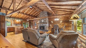 A log cabin interior with a stone fireplace, wooden furniture, and large windows.