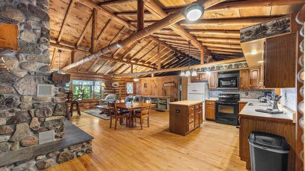 A rustic cabin interior with a stone fireplace, wooden dining table, and a kitchen area with wooden cabinets and appliances.