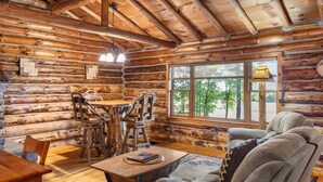 A log cabin interior with a wooden dining table, chairs, a sofa, and a large window offering a view of trees.