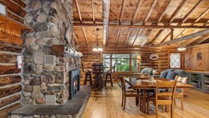 A rustic cabin interior with a stone fireplace, wooden dining table, and chairs.