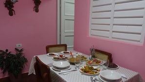 Mesa de jantar posta com diversos pratos, rodeada por cadeiras, num quarto com paredes cor-de-rosa e porta branca.