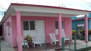 Um edifício rosa e branco com varanda coberta, cadeiras de balanço brancas e um pátio cercado.