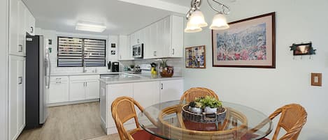 A modern kitchen with white cabinets, a glass dining table, wicker chairs, and a hanging light fixture.