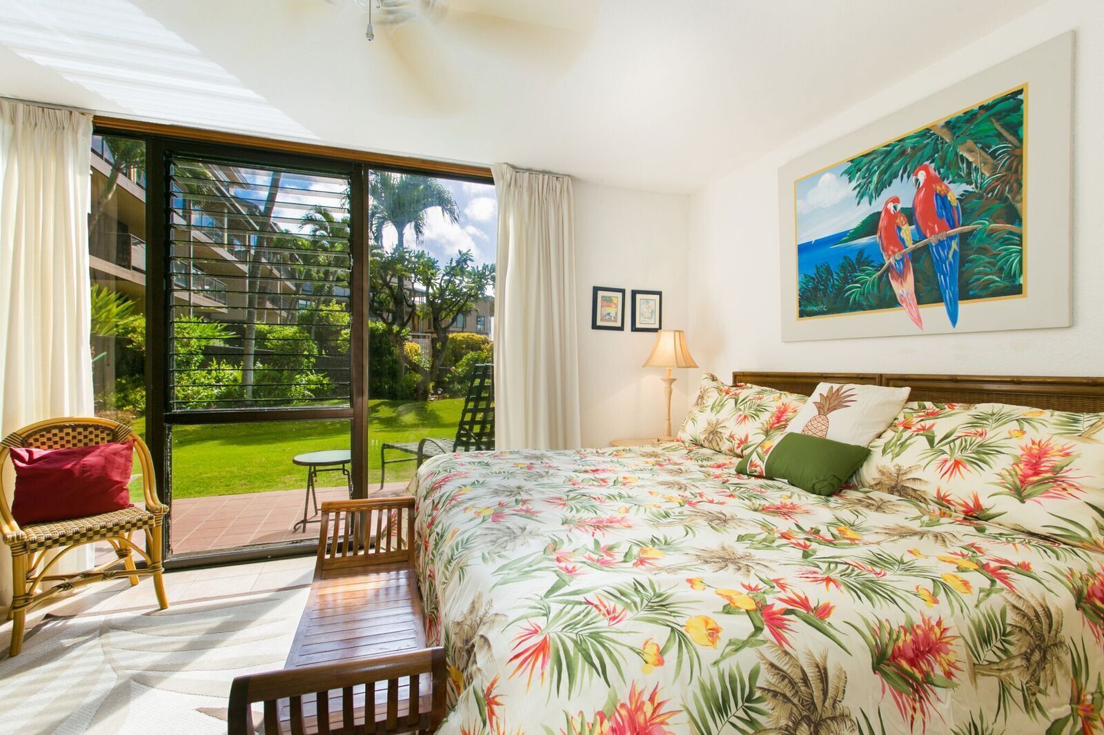 A hotel room with a bed, a chair, a table, and a view of a garden and building through a large window.