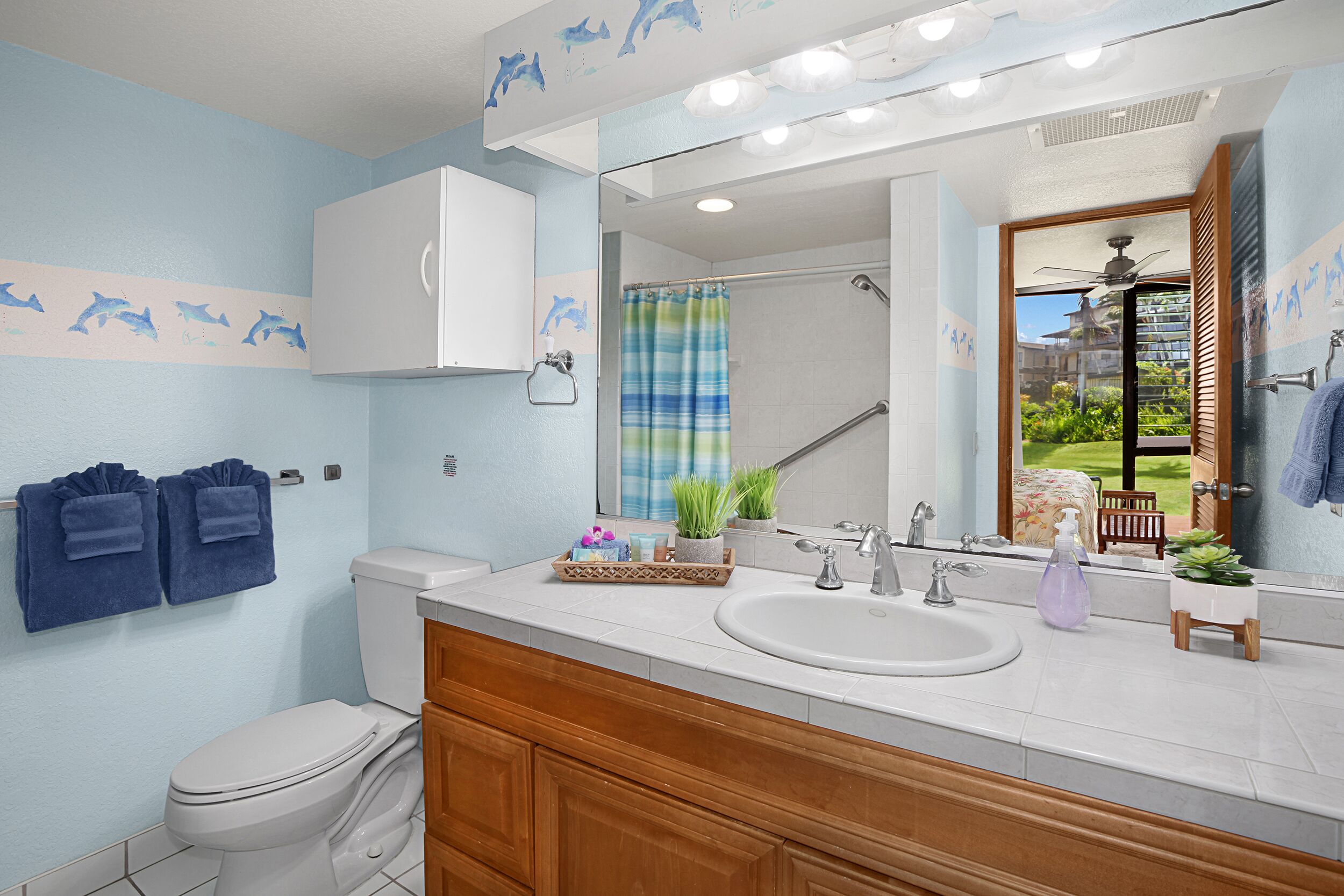 Une salle de bain avec un lavabo blanc, un miroir, une douche avec un rideau bleu et des toilettes. Deux serviettes sont accrochées au mur et une petite plante est posée sur le comptoir du lavabo.
