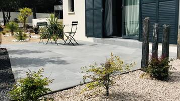 Un edificio moderno con una puerta de garaje, un patio con mesa y sillas, y una zona de grava con pequeñas plantas.