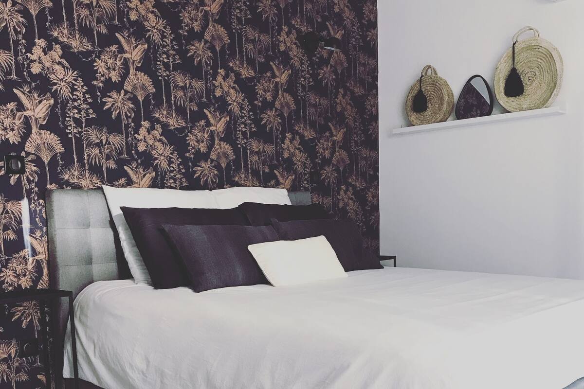 A neatly made bed with a white duvet and dark pillows, a dark headboard, and a wall with a tropical leaf pattern. There are two woven baskets on a shelf above the bed.