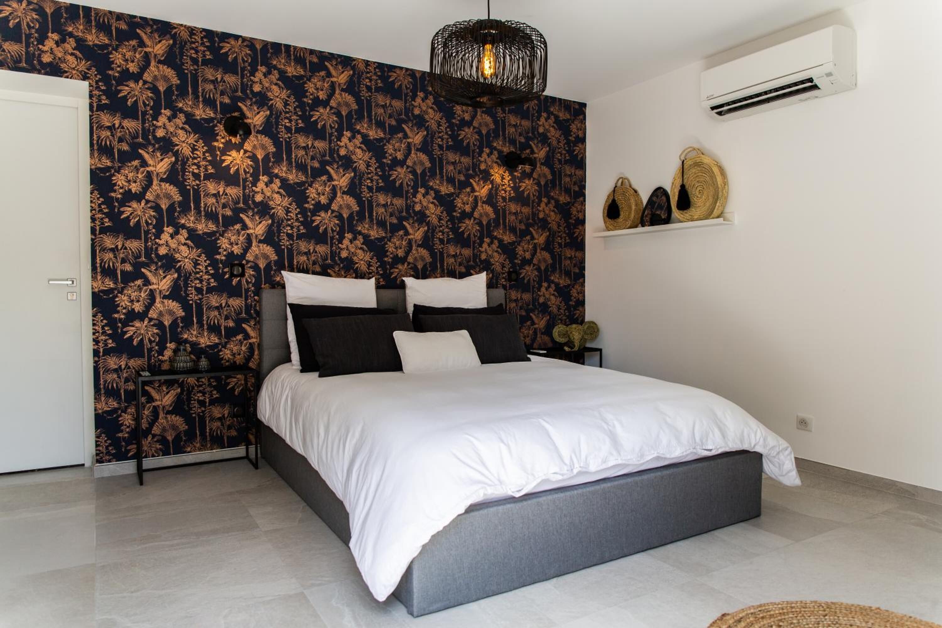 Una camera da letto con un letto, carta da parati a motivi scuri, una lampada a soffitto bianca, un condizionatore d'aria e uno scaffale con oggetti decorativi.