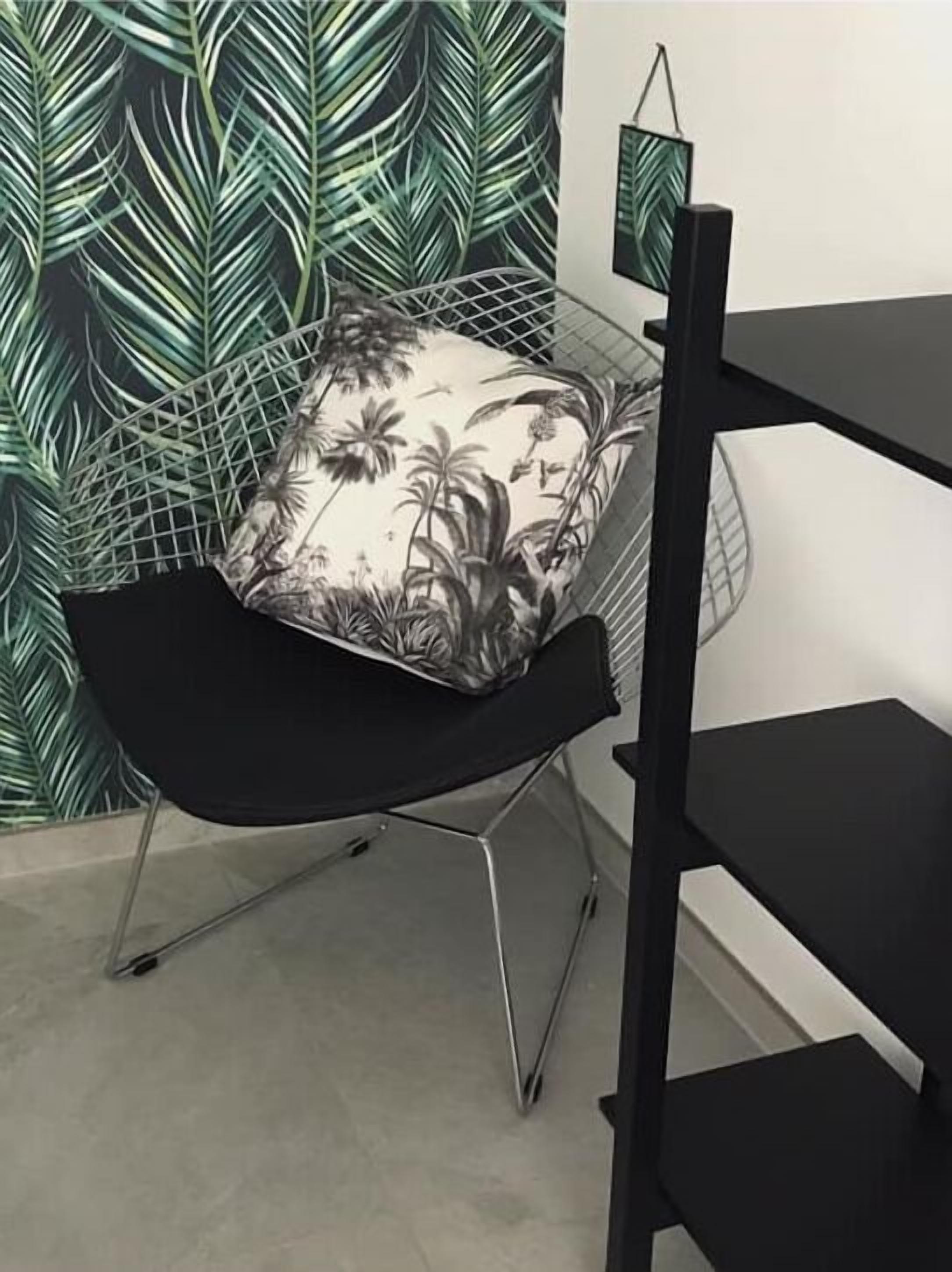 Ett rum med en svart metallstol, en vit kudde med ett tropiskt mönster och en vägg med ett grönt bladmotiv.