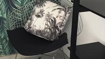 Ett rum med en svart metallstol, en vit kudde med ett tropiskt mönster och en vägg med ett grönt bladmotiv.