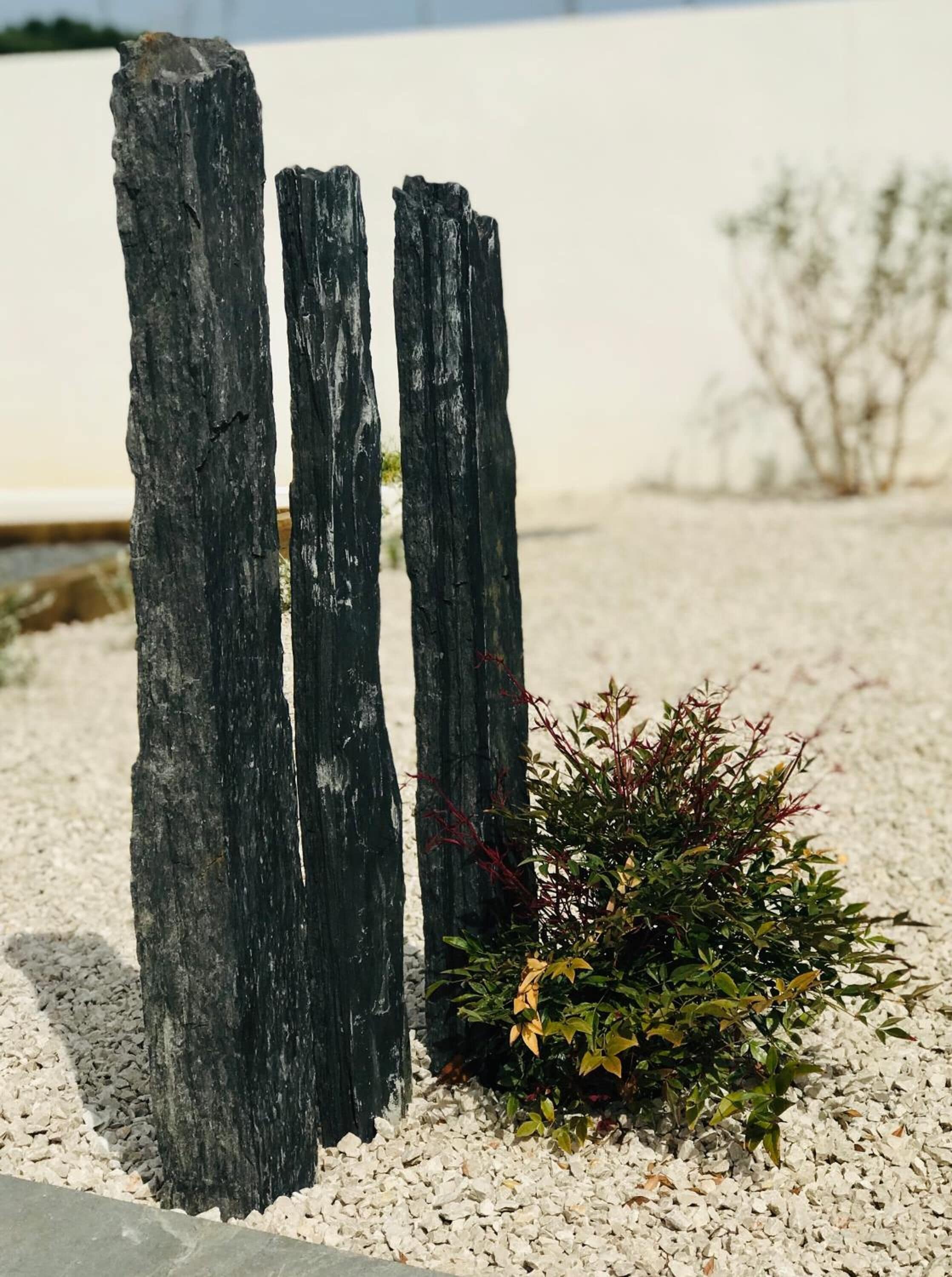 Trois poteaux en bois verticaux, plantés dans un espace recouvert de gravier, avec un petit buisson à proximité.