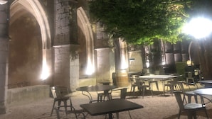 Un'area pranzo all'aperto con tavoli e sedie, archi in pietra e un albero che offre ombra.
