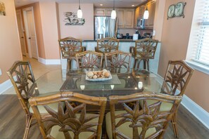 Un coin repas avec une table en verre entourée de six chaises en osier, un élément décoratif mural au-dessus de la table, et une cuisine en arrière-plan.