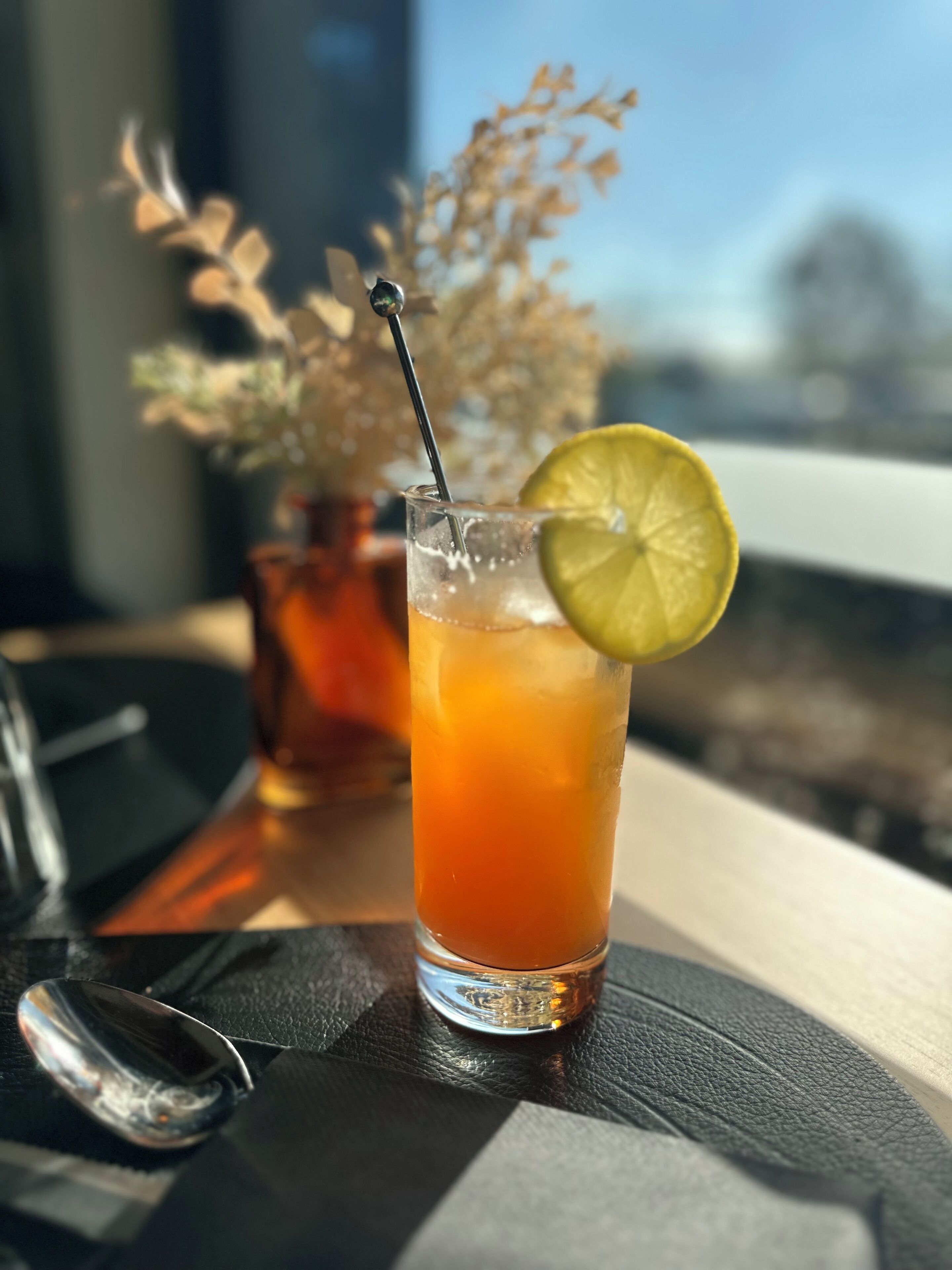 Un bicchiere alto con una cannuccia, contenente una bevanda, guarnito con una fetta di lime, su un tavolo con un cucchiaio e un vaso di fiori secchi sullo sfondo.