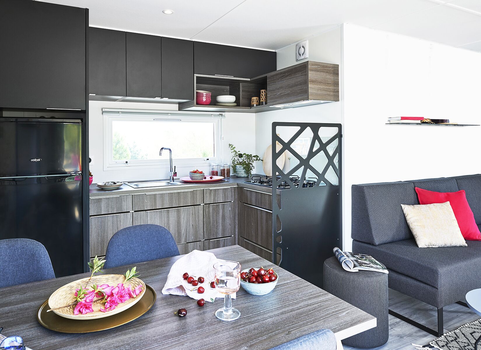 A modern kitchen with a built-in refrigerator, a dining table with a bowl of fruit and a glass, and a grey sofa with red cushions.