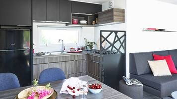 A modern kitchen with a built-in refrigerator, a dining table with a bowl of fruit and a glass, and a grey sofa with red cushions.