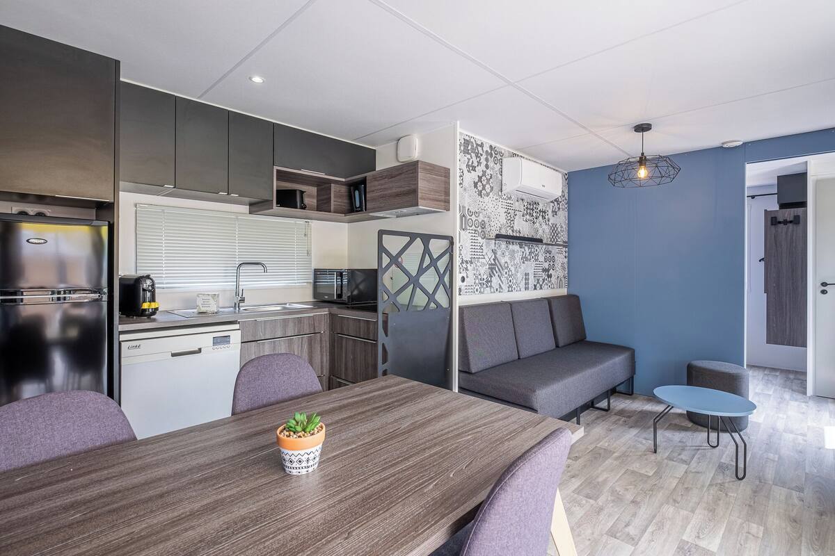 Une cuisine moderne équipée d’un réfrigérateur, d’un four à micro-ondes et d’un évier. On y trouve une table à manger avec des chaises et une petite plante en pot. Un canapé gris et une chaise bleue sont également visibles.