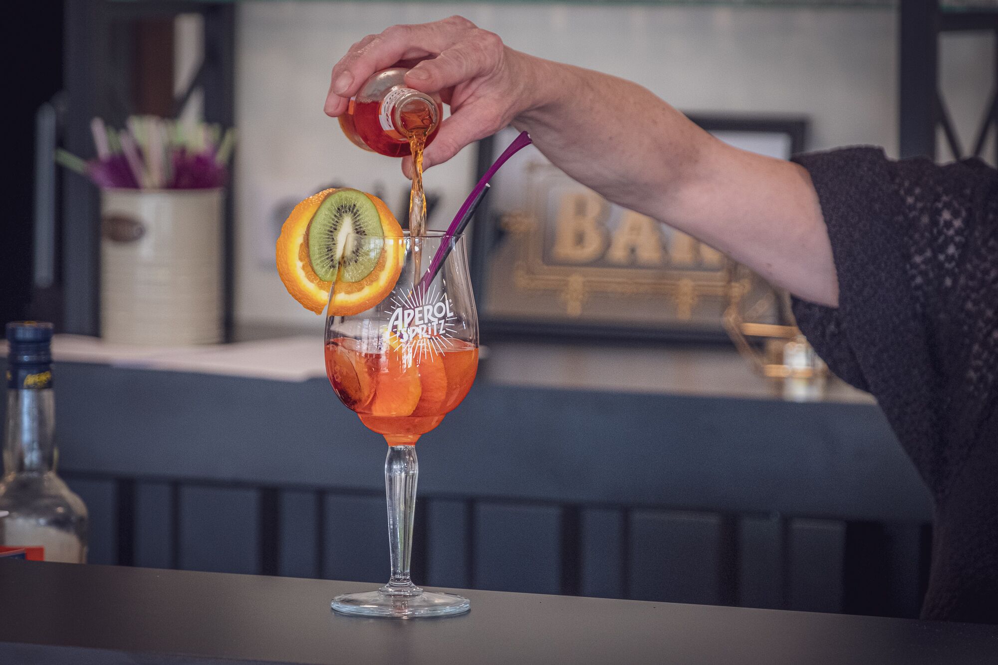 Un cantinero sirve una bebida en un vaso con pajita, decorada con una rodaja de naranja y un kiwi.