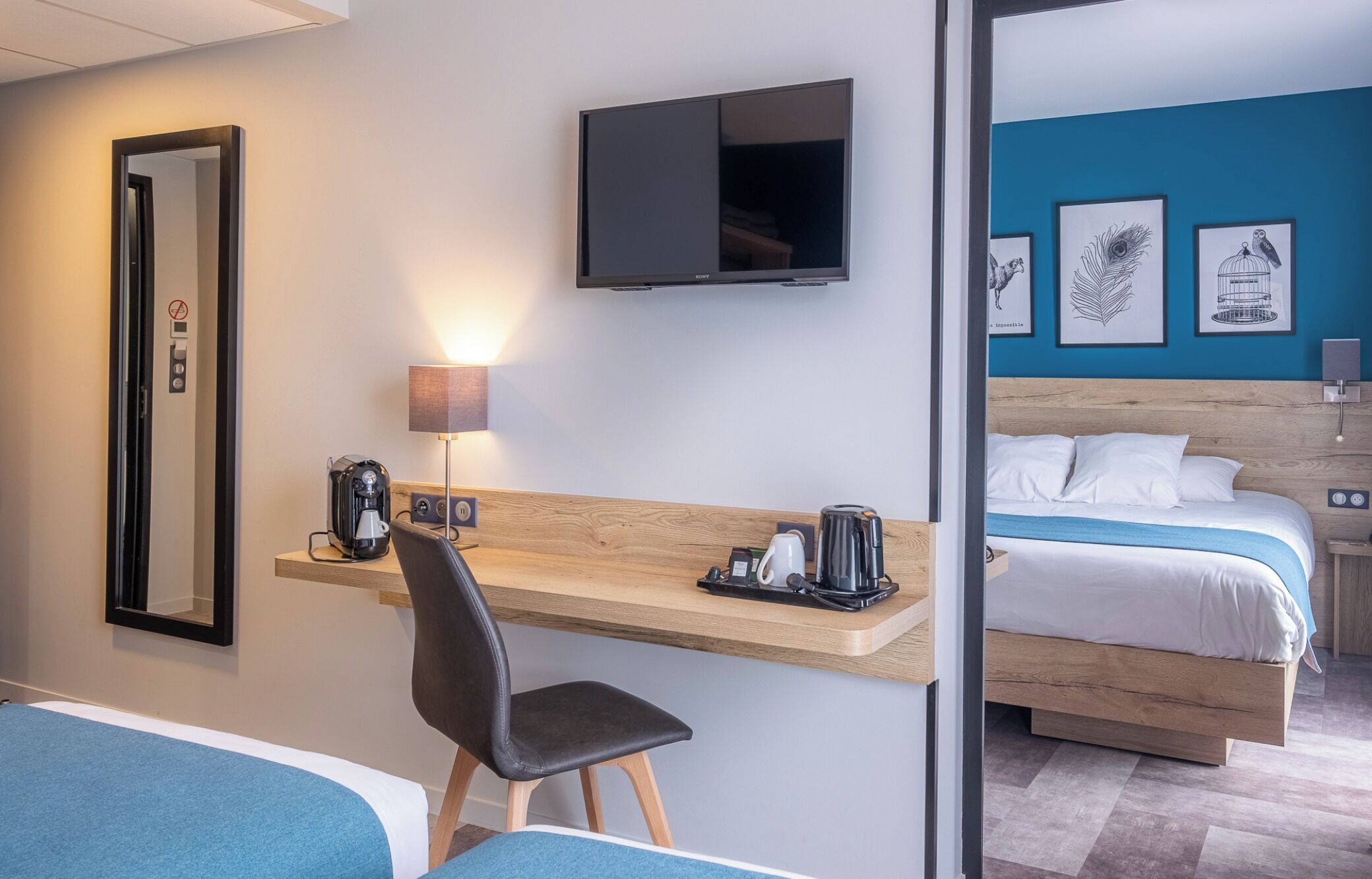 A hotel room with a desk, chair, bed, TV, and artwork on the wall.