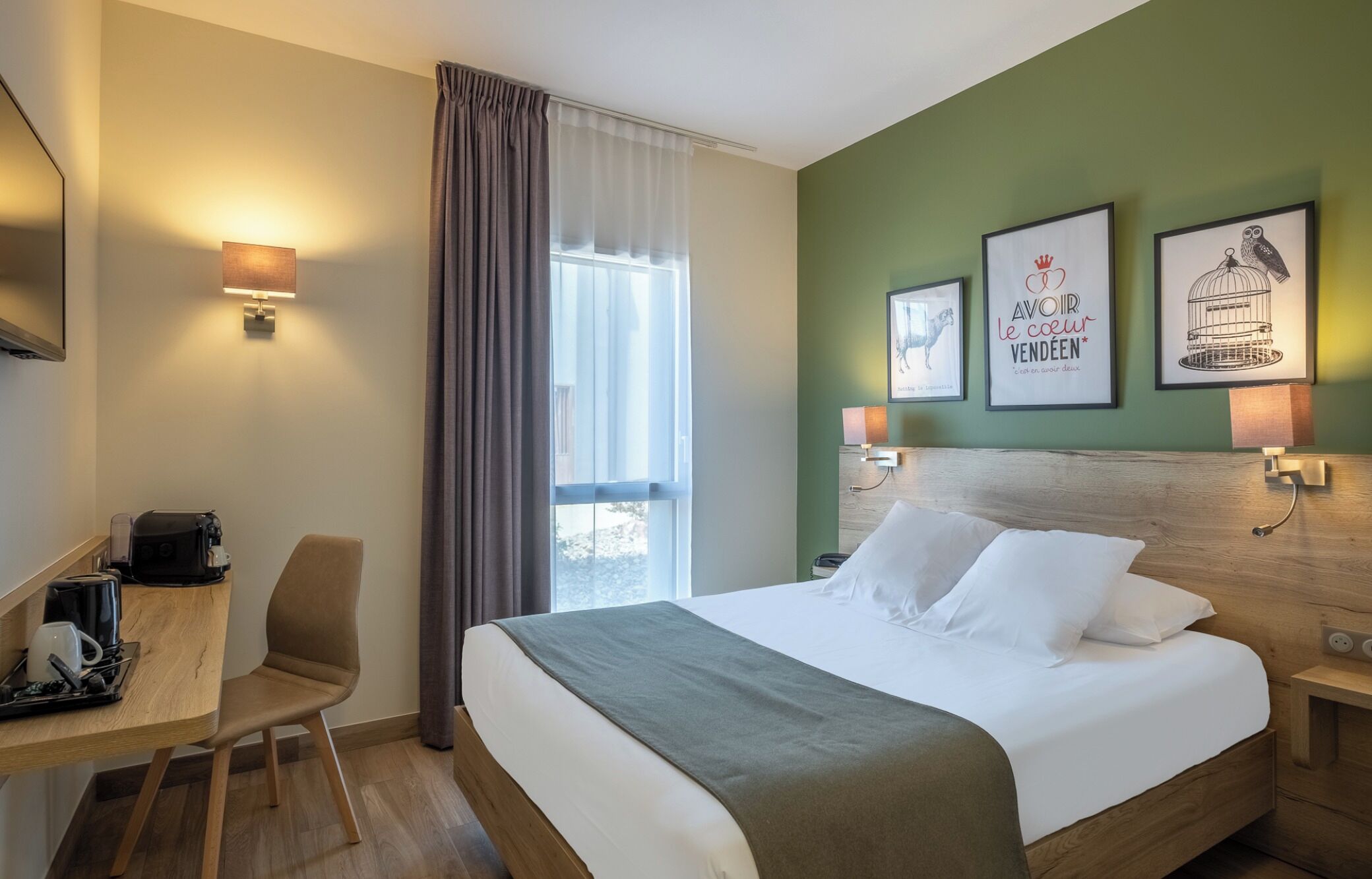 A hotel room with a bed, a desk with a coffee machine, a chair, a window with curtains, and wall art.