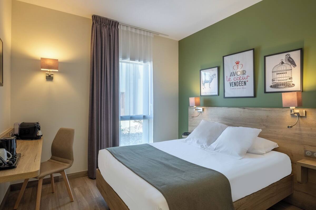 Une chambre d’hôtel comprenant un lit, un bureau avec une machine à café, une chaise, une fenêtre avec des rideaux et des œuvres d’art murales.