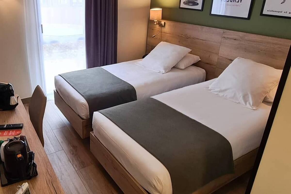 Une chambre d’hôtel avec deux lits, un bureau avec une chaise, une lampe et une fenêtre avec des rideaux.