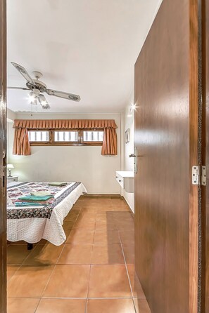 A bedroom with a bed, a ceiling fan, a window with curtains, a bedside table, and a wooden door. - 1 bdr apartment with heated pool, close to the beach, Complex Garden City  (Costa Adeje)