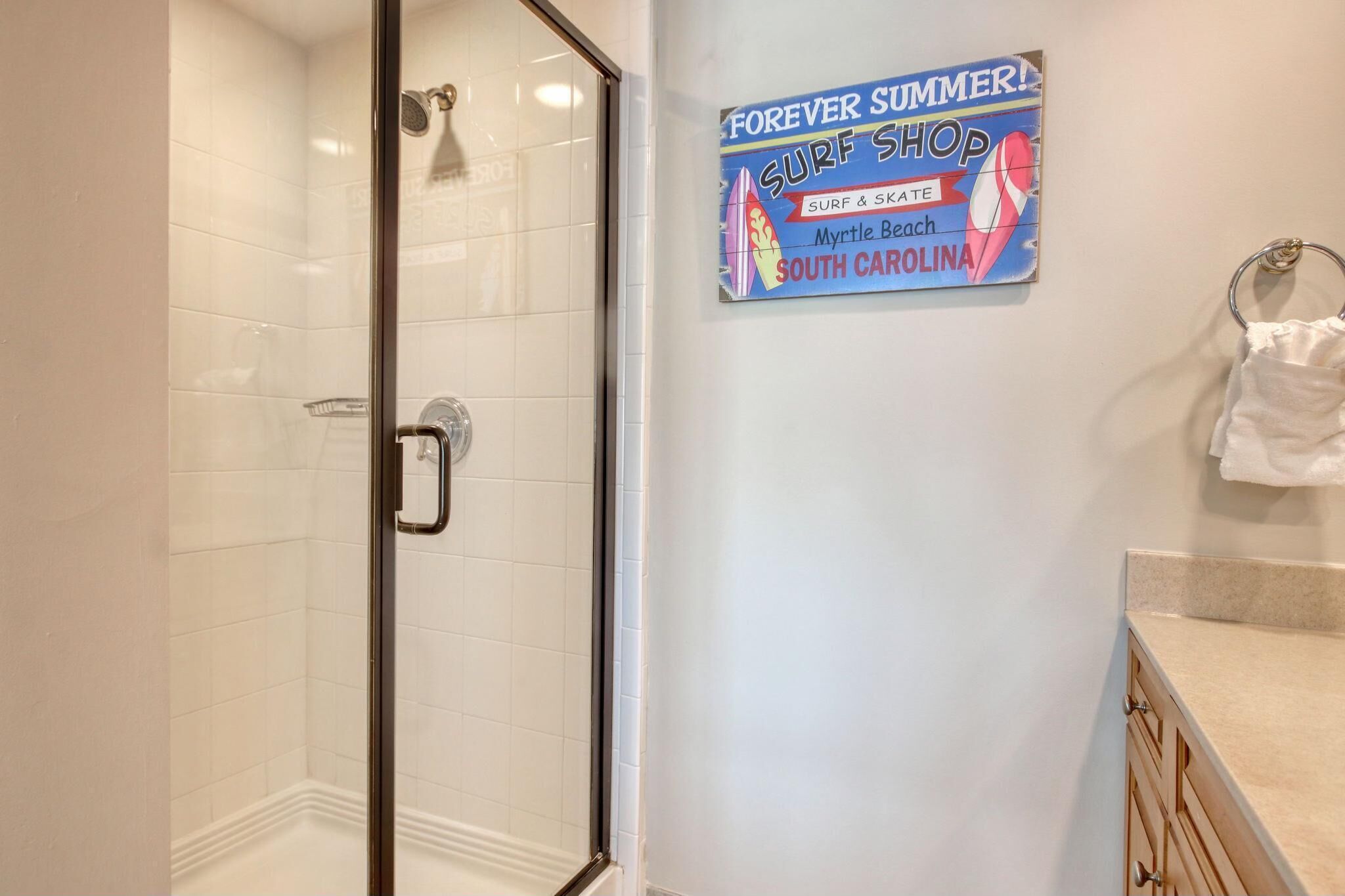 Une salle de bain avec une porte de douche en verre, des carreaux blancs, un porte-serviettes et une affiche publicitaire pour un magasin de surf situé à Myrtle Beach, en Caroline du Sud.