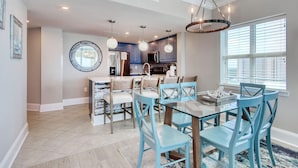 A modern kitchen with a central island, pendant lighting, and a dining area with blue chairs and a glass table. - MVG 1404 Oceanfront Corner Penthouse (North Myrtle Beach)