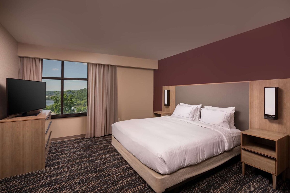 Ein Hotelzimmer mit einem Bett, einem Fernseher, einem Schreibtisch und einem Fenster mit Vorhängen.