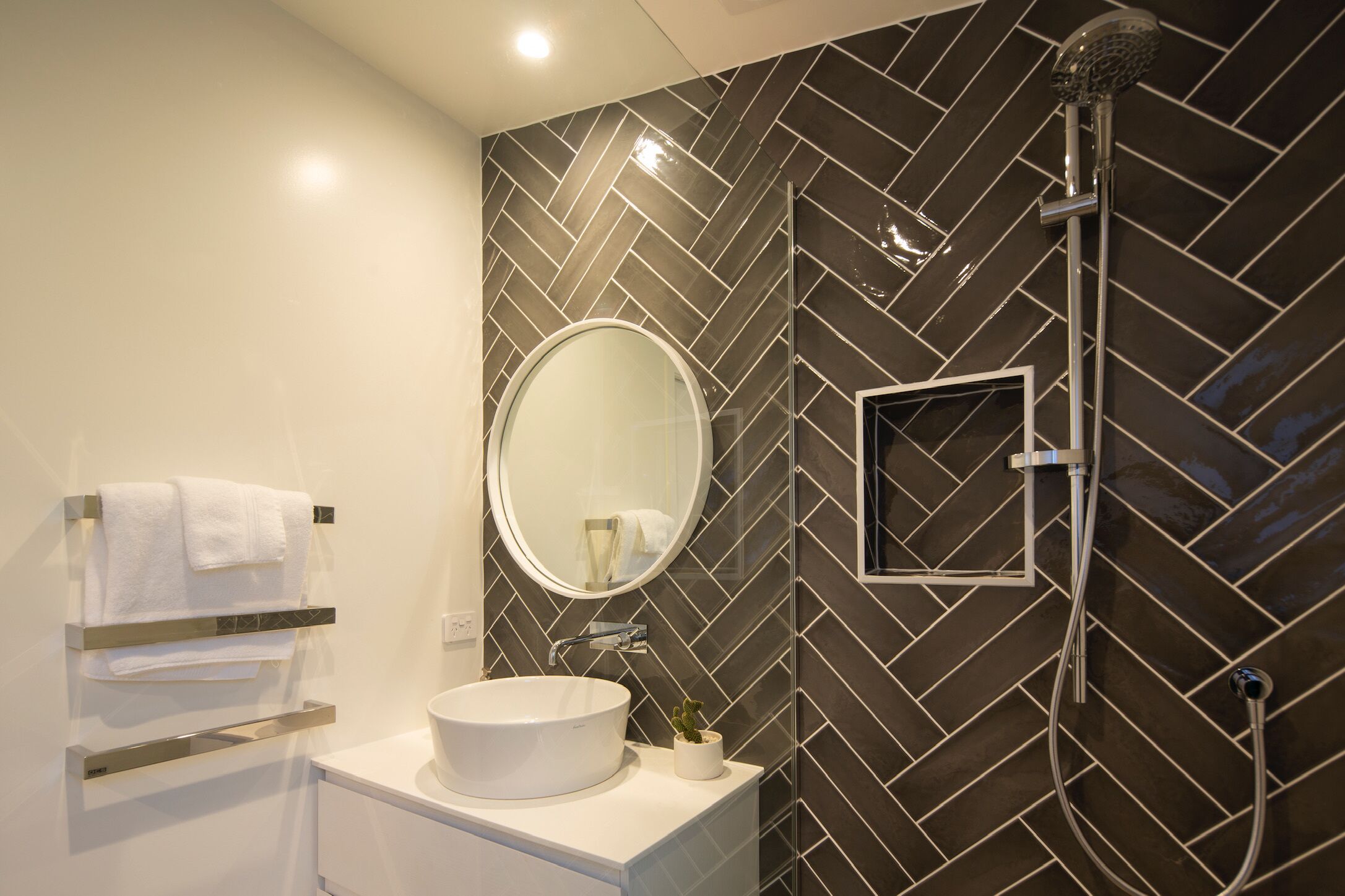 A modern bathroom with a herringbone-patterned wall, a round mirror, a white sink, and a shower with a handheld showerhead.